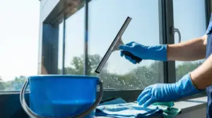 Persona con guanti blu che pulisce una finestra con tergivetro e secchio